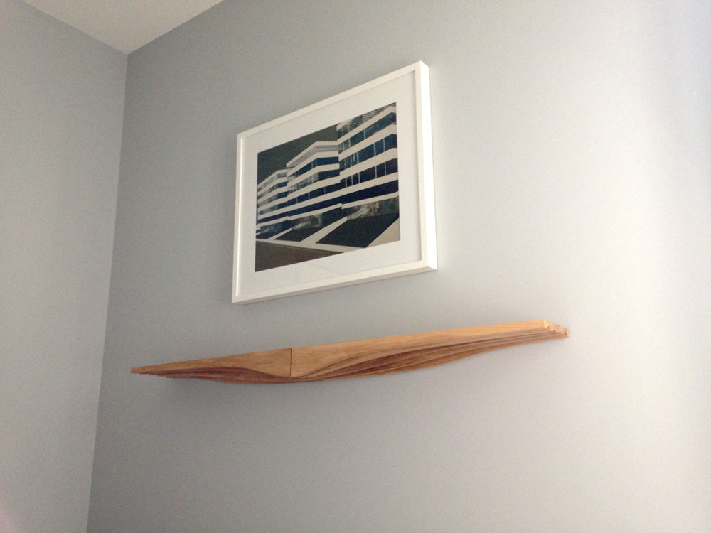 A pair of natural wood coloured contour shelves holding nothing but the air above them, with a framed print hung above them on a grey wall.